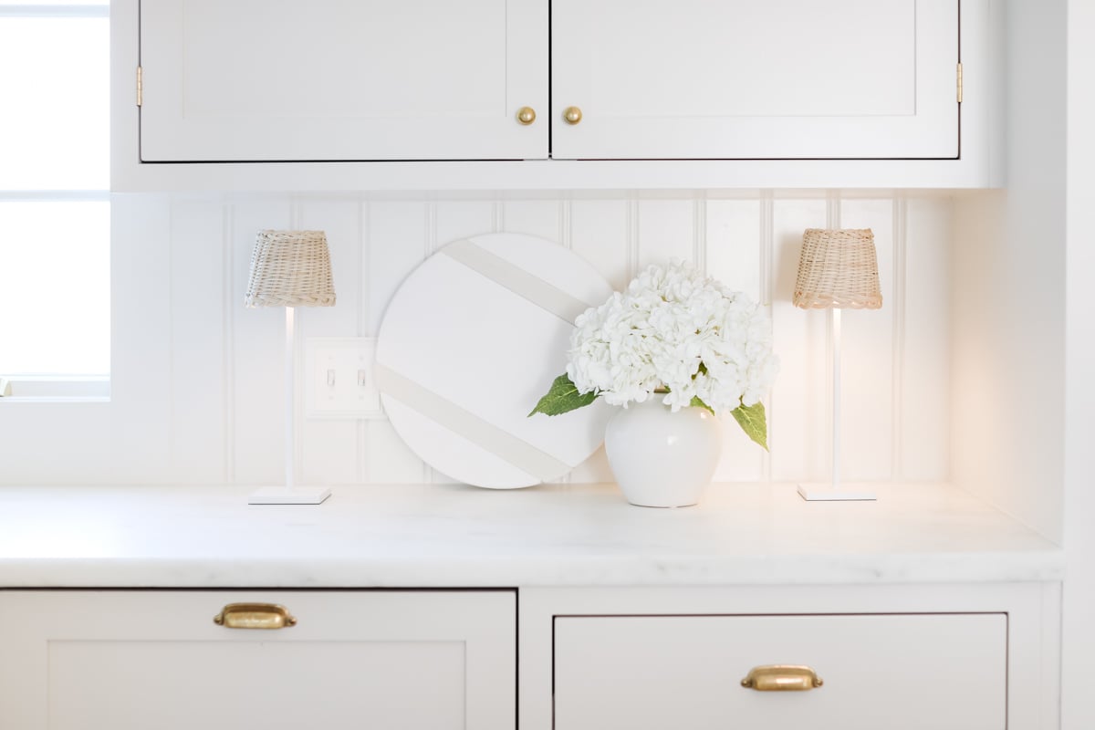 A minimalist kitchen interior with white cabinets featuring precise shaker cabinet knob placement, marble countertop, two small lamps, and a vase with white hydrangeas.