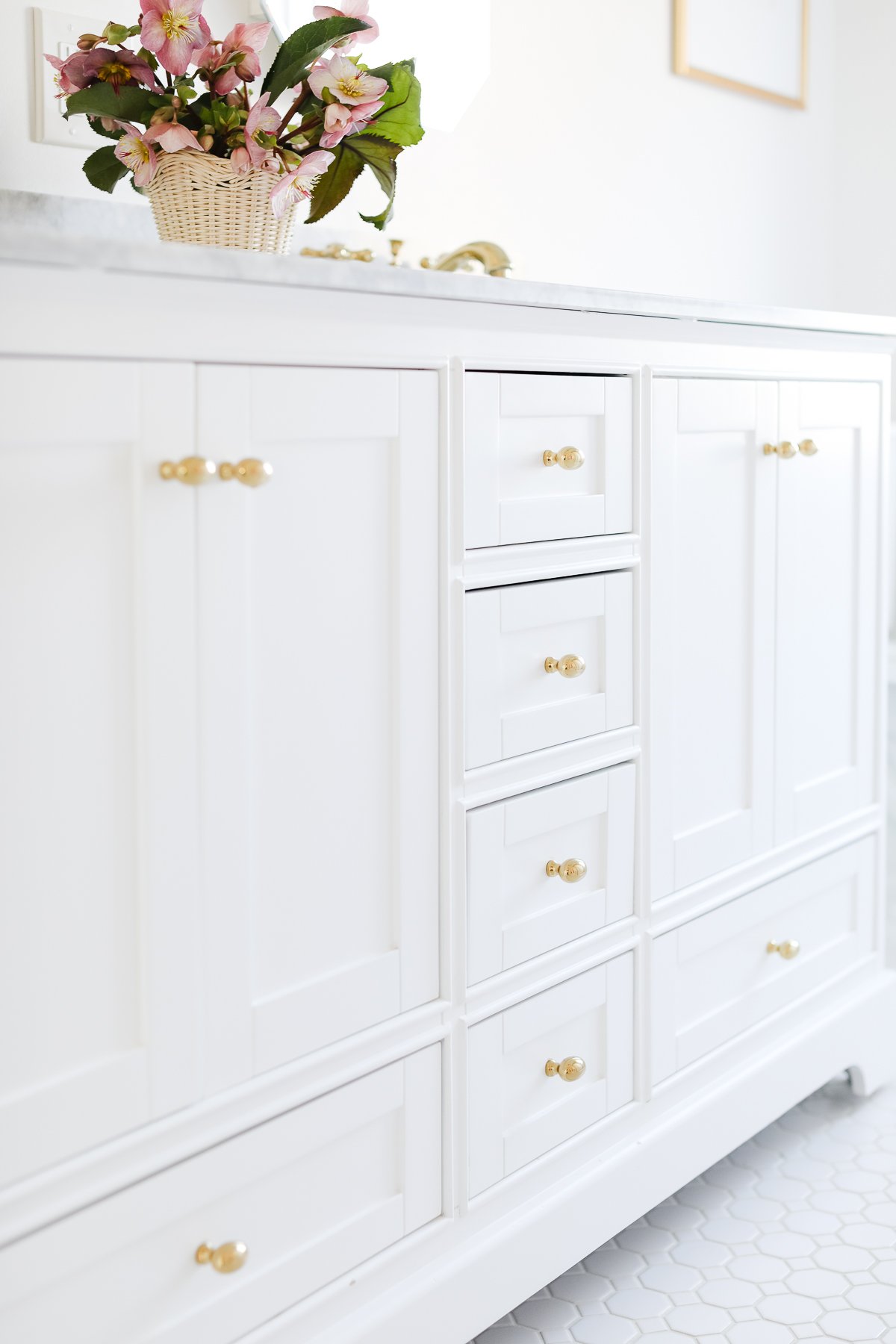 White bathroom vanity with gold handles, showcasing precise cabinet knob placement, and a basket of flowers on top.