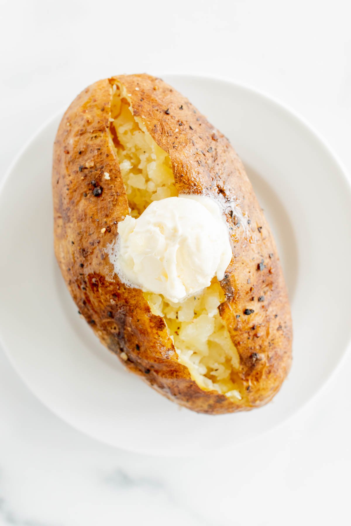 A grilled baked potato on a white plate with a topping of butter and sour cream.