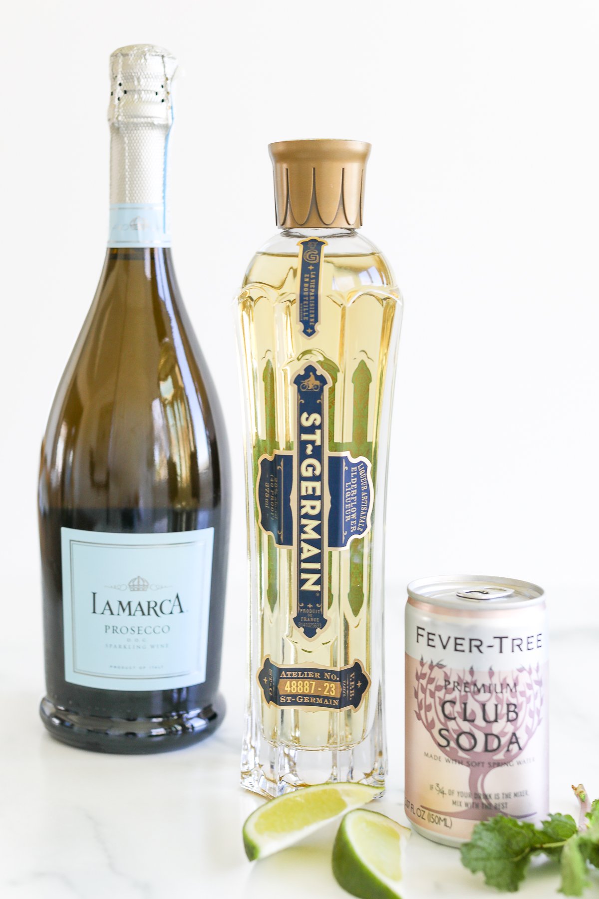 A bottle of St-Germain elderflower liqueur, a bottle of LaMarca prosecco, a can of Fever-Tree premium club soda, and slices of lime arranged on a marble surface for