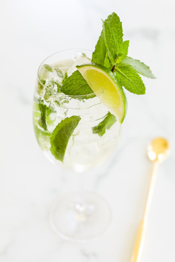 A glass of Hugo spritz with mint leaves and a lime wedge, accompanied by a gold-colored stirring spoon on a marble surface.
