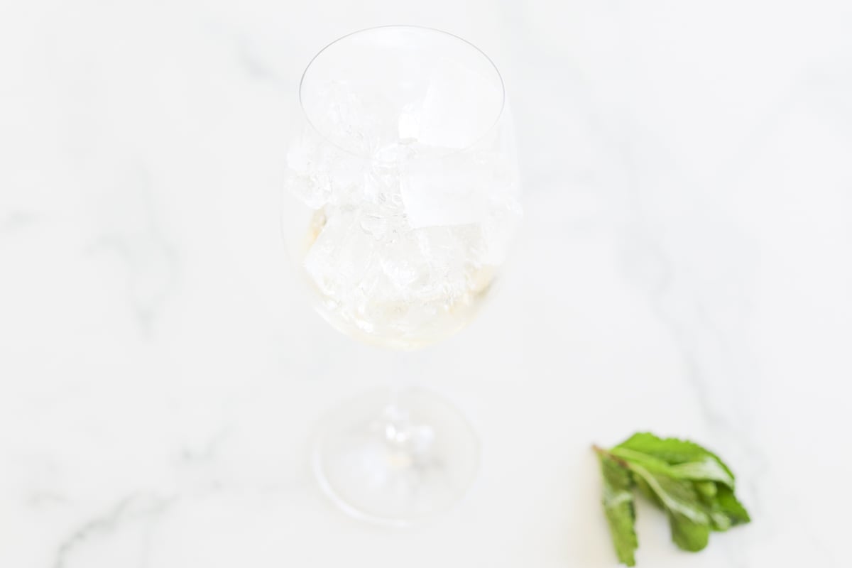 A wine glass filled with ice cubes on a marble surface with a sprig of mint leaves nearby.