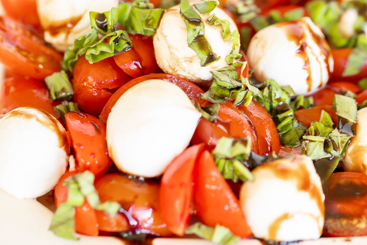 A close-up of an Italian caprese salad with sliced tomatoes, mozzarella cheese, and chopped basil, drizzled with balsamic glaze.