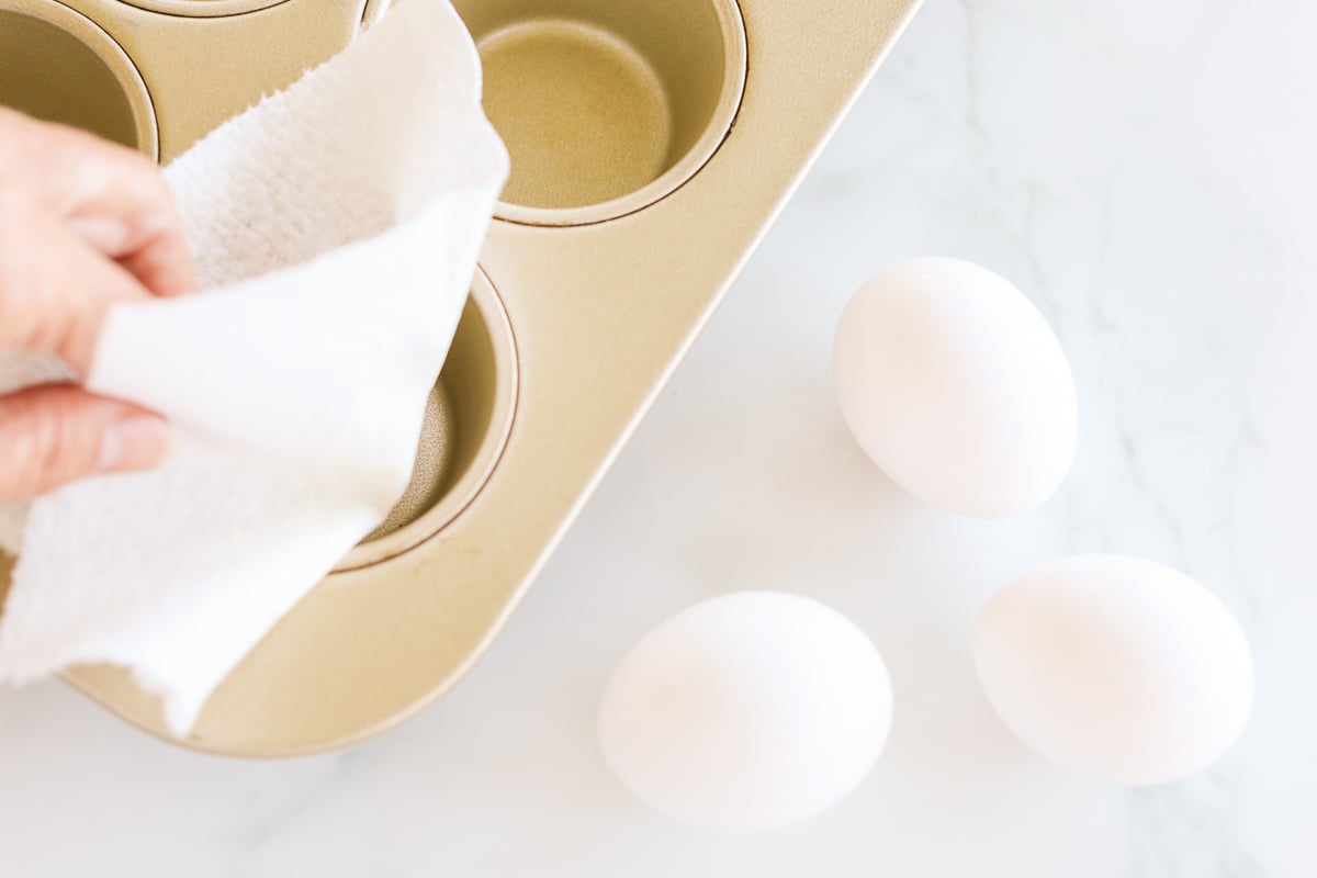 Greasing a muffin tin next to three white eggs on a marble countertop.