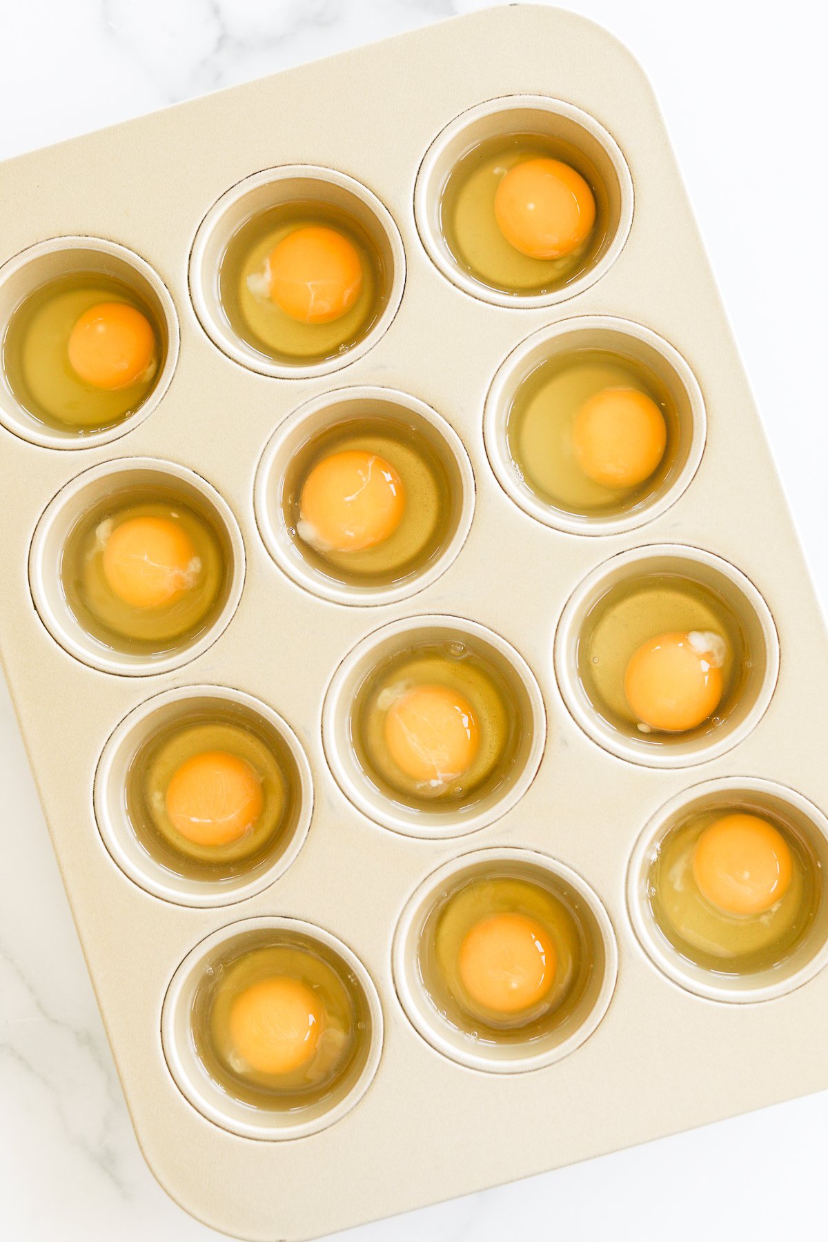 A dozen raw eggs cracked into a muffin tin, ready for baking or cooking.