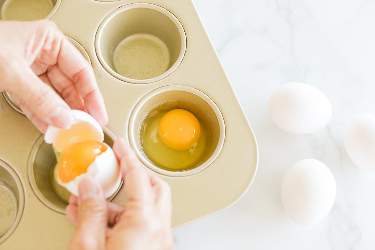 Cracking an egg into a muffin tin with whole eggs beside it.