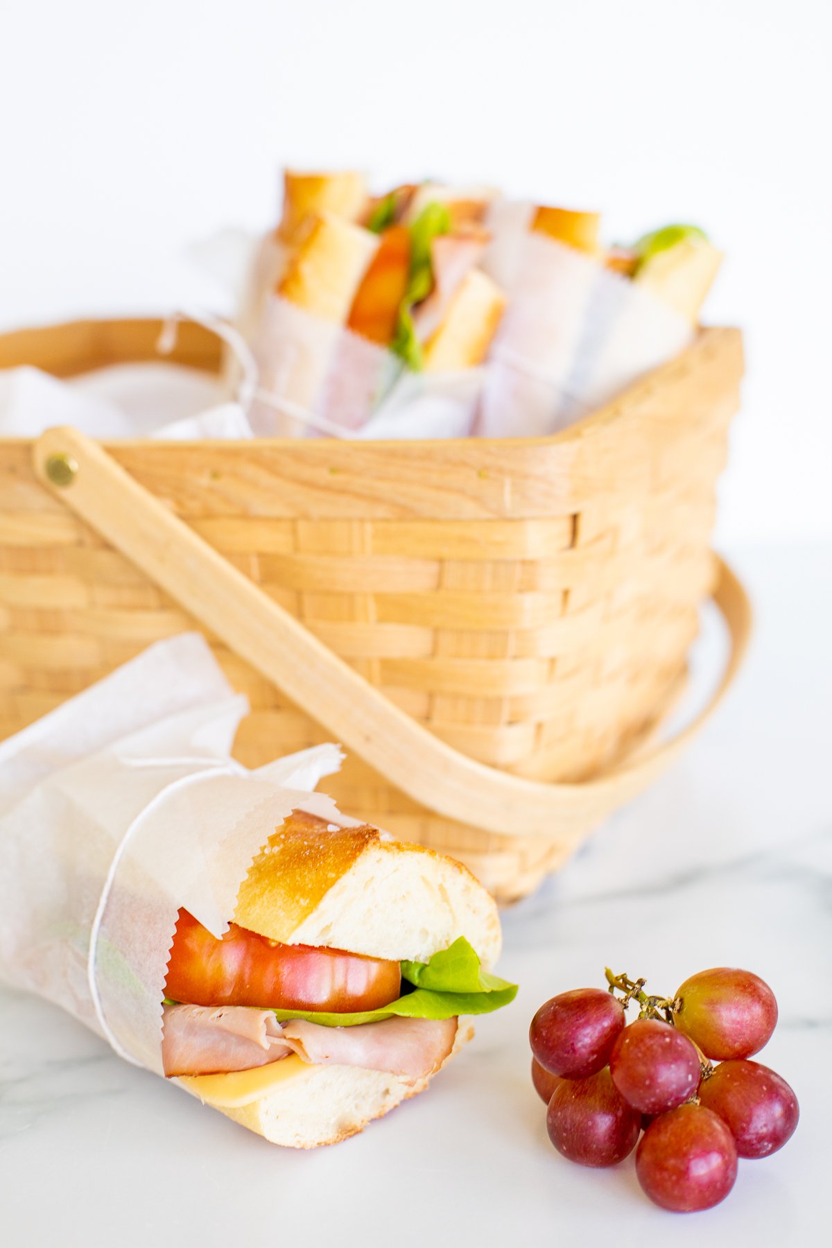 A picnic basket with wrapped sandwiches and a bunch of red grapes on a white surface.