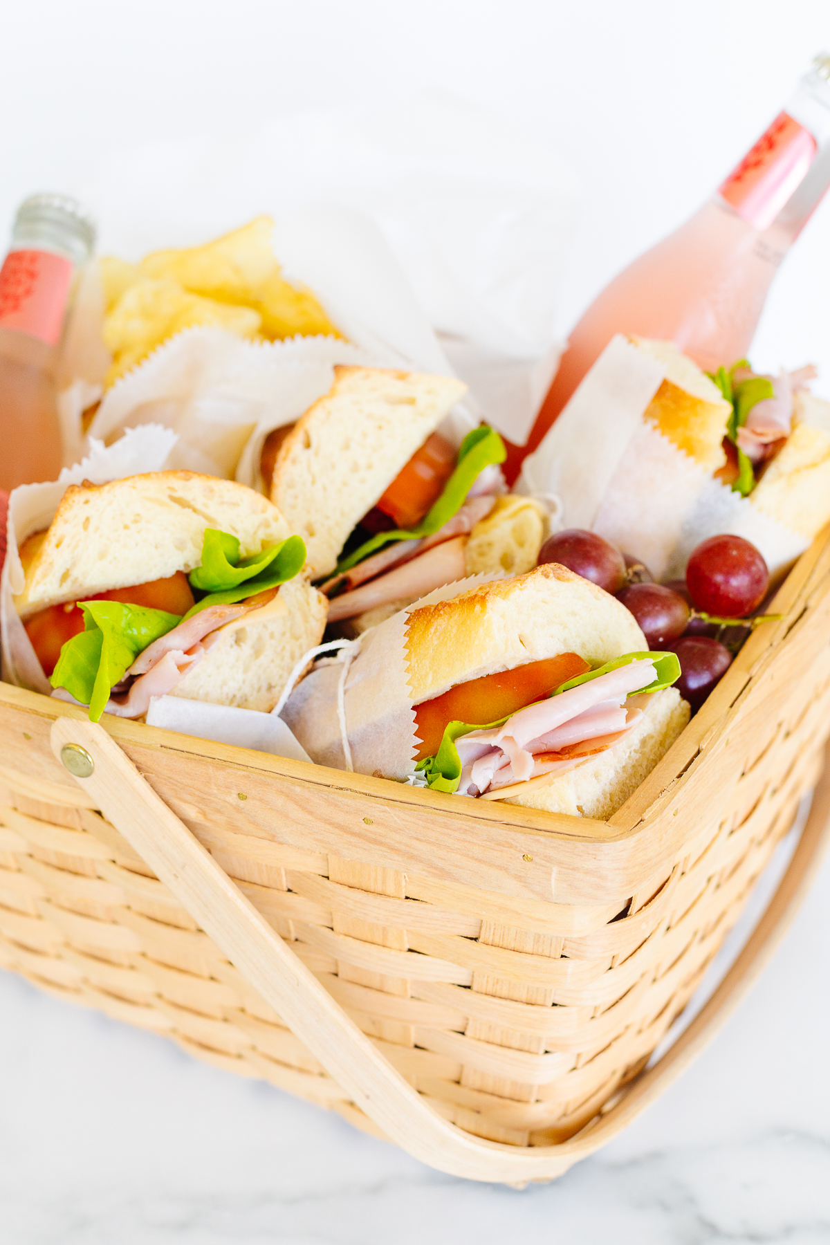 A picnic basket filled with sandwiches, chips, grapes, and a bottle of beverage.