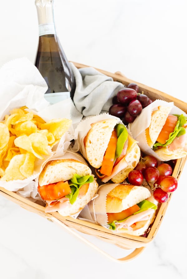 A picnic basket containing sandwiches, chips, grapes, and a bottle of sparkling beverage.