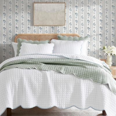 A neatly made bed with white and light green scalloped bedding, a cane headboard, floral wallpaper, and a vase of flowers on a bedside table creates a charming space accented with scalloped decor.