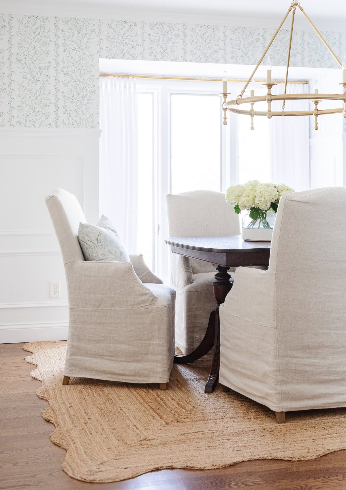 A dining area with four beige upholstered armchairs around a dark wooden table, a woven rug, floral wallpaper, scalloped decor accents, and a brass chandelier overhead.
