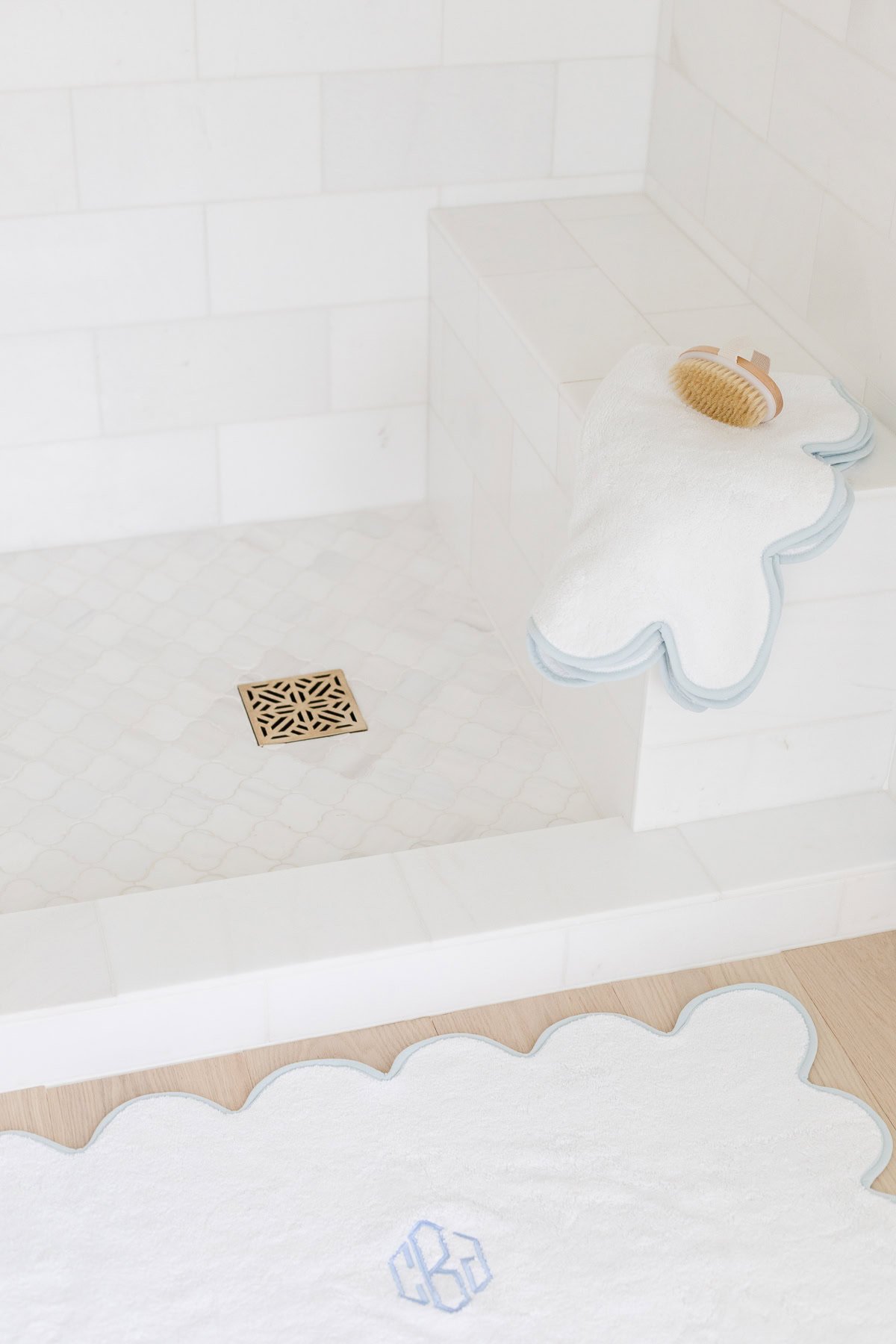 A white tiled shower with a gold drain, featuring a white bath mat on the floor and a white towel with a cracked egg design on the step adds playful charm to this scalloped decor-inspired space.