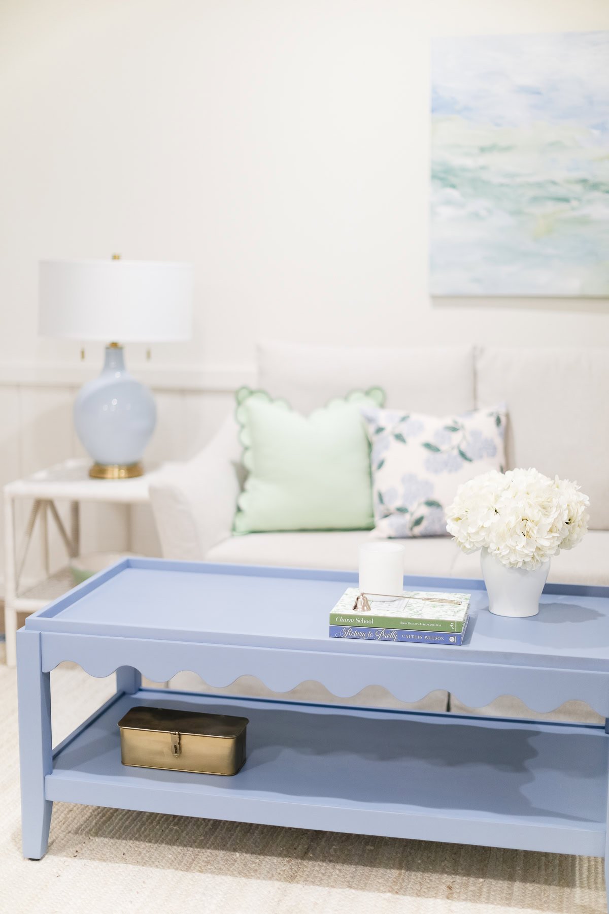 A light blue coffee table with books, a vase of white flowers, and a tissue box sits in front of a white couch with pastel pillows; scalloped decor and wall art add charm to the cozy setting.