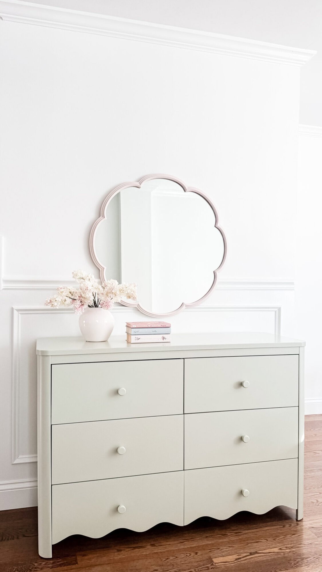A light-colored dresser with six drawers stands against a white wall, topped with a vase of flowers, two stacked books, and a decorative round mirror above it, adding subtle scalloped decor charm to the space.