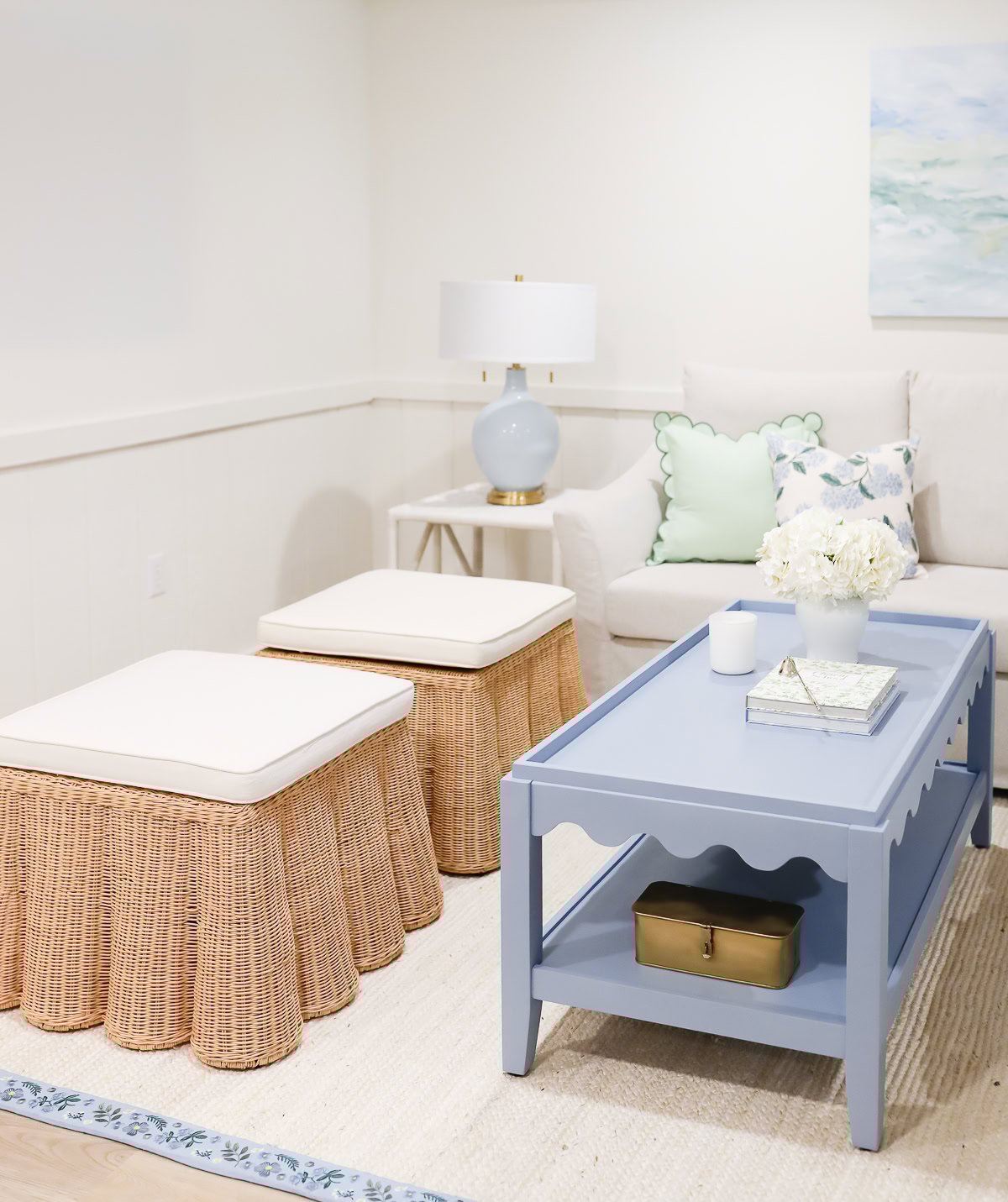A bright living room with a white sofa, light blue coffee table, two wicker ottomans, and scalloped decor touches like a blue lamp on the side table and a vase of white flowers on the table.