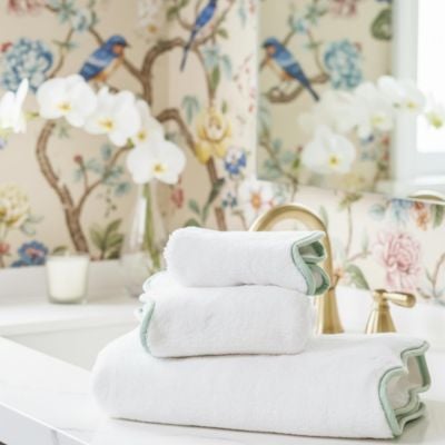 Three neatly folded white towels with light green scalloped decor trim are stacked on a bathroom counter next to a gold faucet, with floral wallpaper and orchids in the background.
