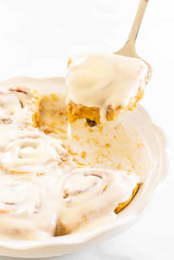 A spoon lifting a cinnamon roll covered in cream cheese icing from a dish with more rolls.