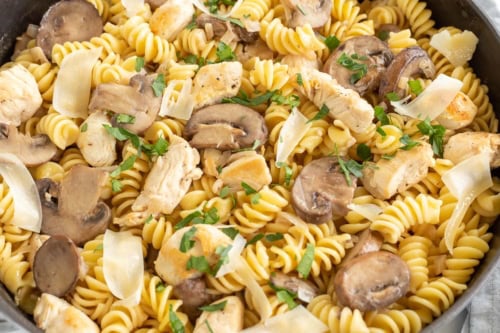 A skillet containing cooked fusilli pasta with chicken, mushrooms, onions, and garnished with chopped parsley.
