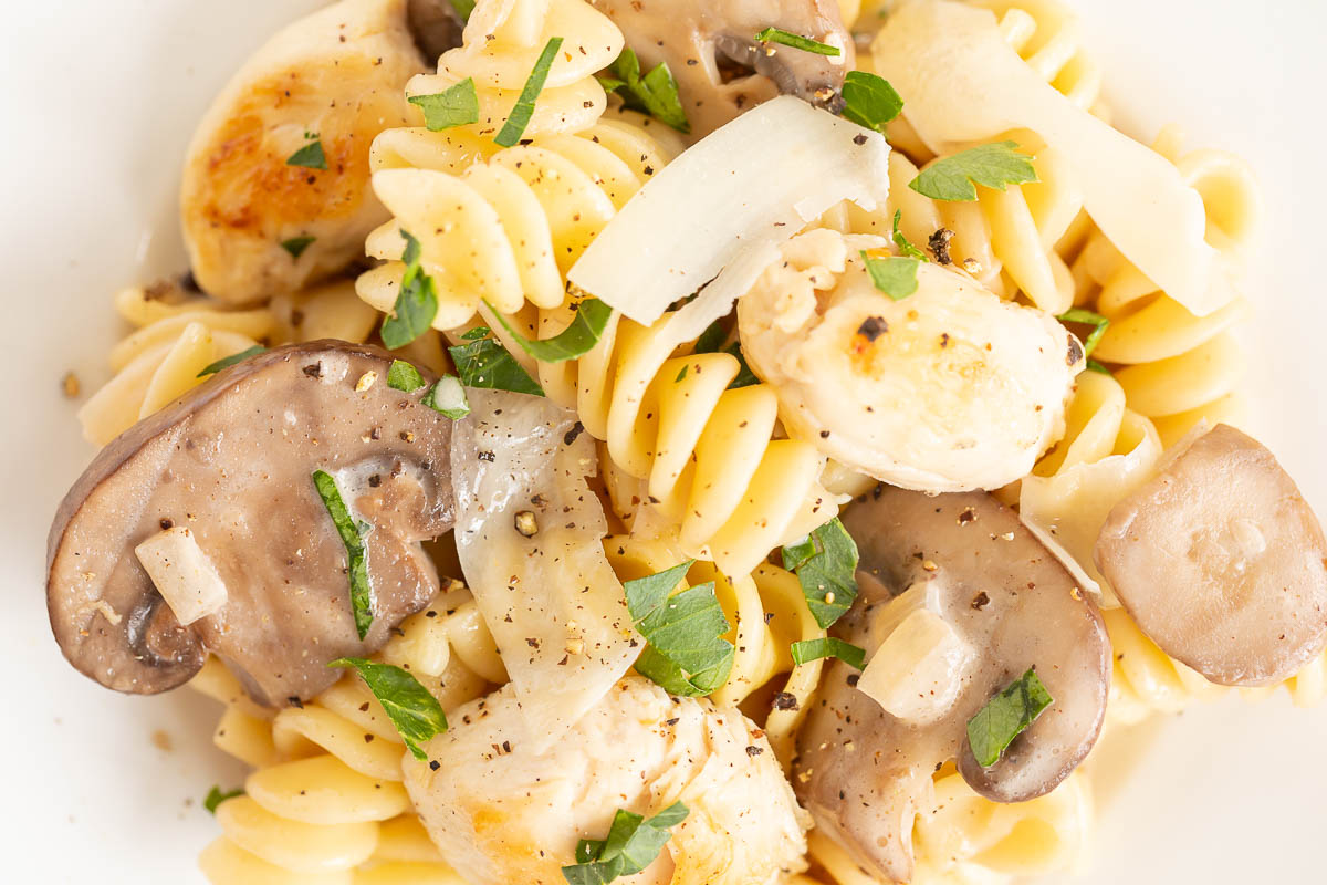 Close-up of a chicken mushroom pasta dish with grilled chicken, sliced mushrooms, and chopped herbs on a white plate.