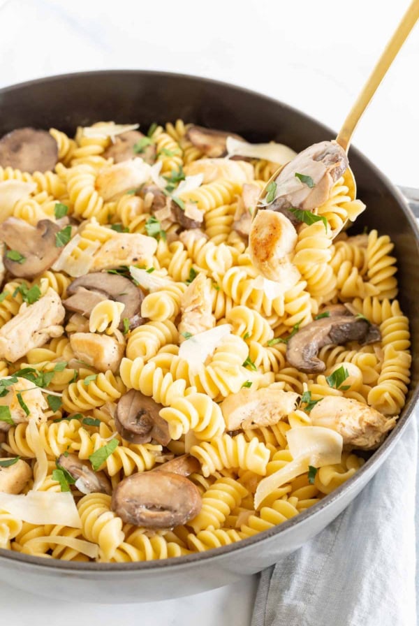A skillet filled with creamy chicken and mushroom pasta, garnished with parsley, with a spoon lifting a portion.