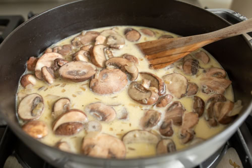 Sliced chicken and mushrooms cooking in a creamy sauce in a skillet, with a wooden spoon.