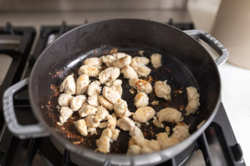 Diced chicken and mushroom cooking in a black skillet on a stove, with visible browning and seasoning.