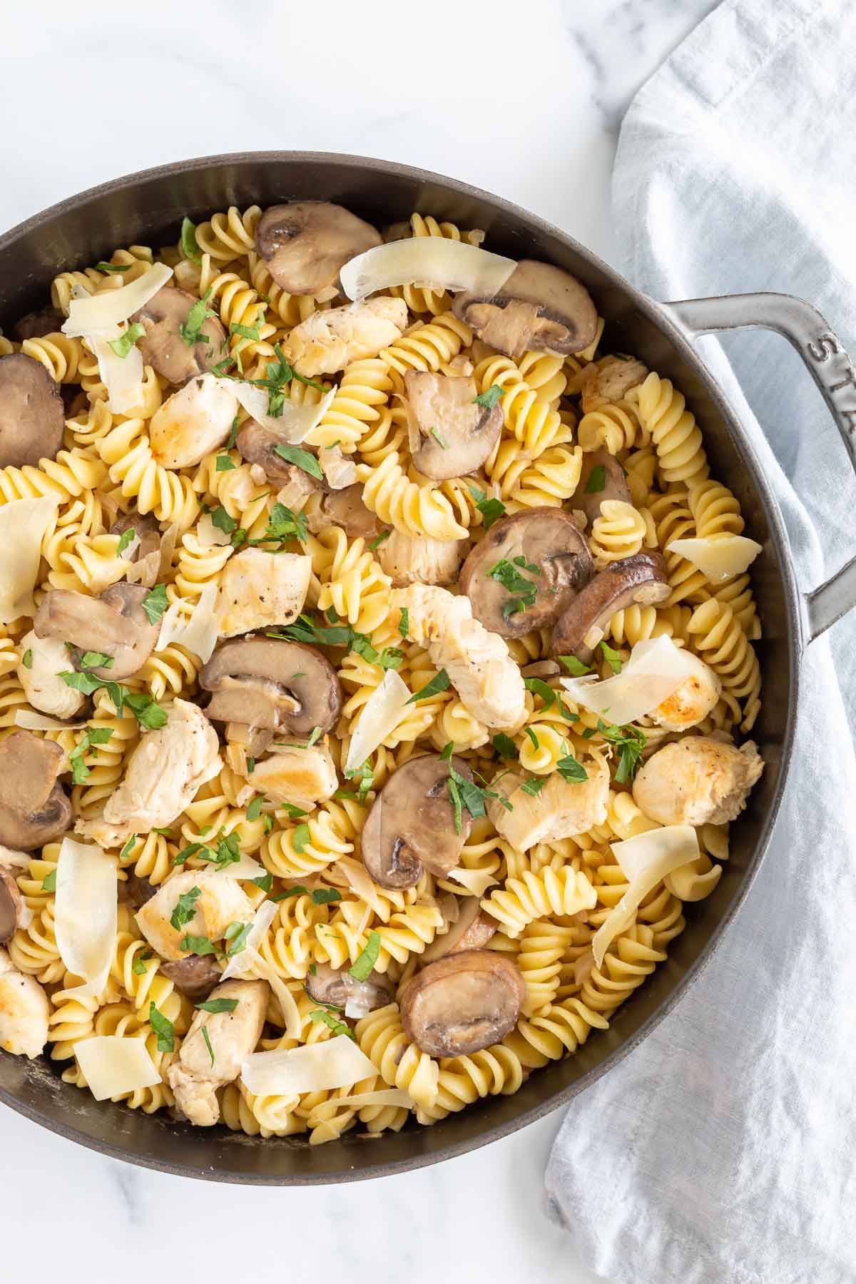 A skillet containing chicken mushroom pasta mixed with fusilli, garnished with herbs on a marble surface.