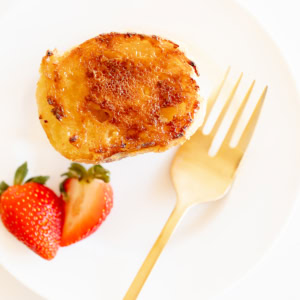 Creme Brulee French toast slice on a white plate with a gold fork. Garnished with sliced strawberries.