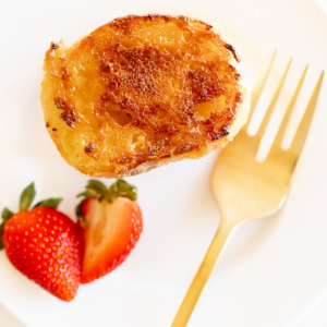 Creme Brulee French toast slice on a white plate with a gold fork. Garnished with sliced strawberries.