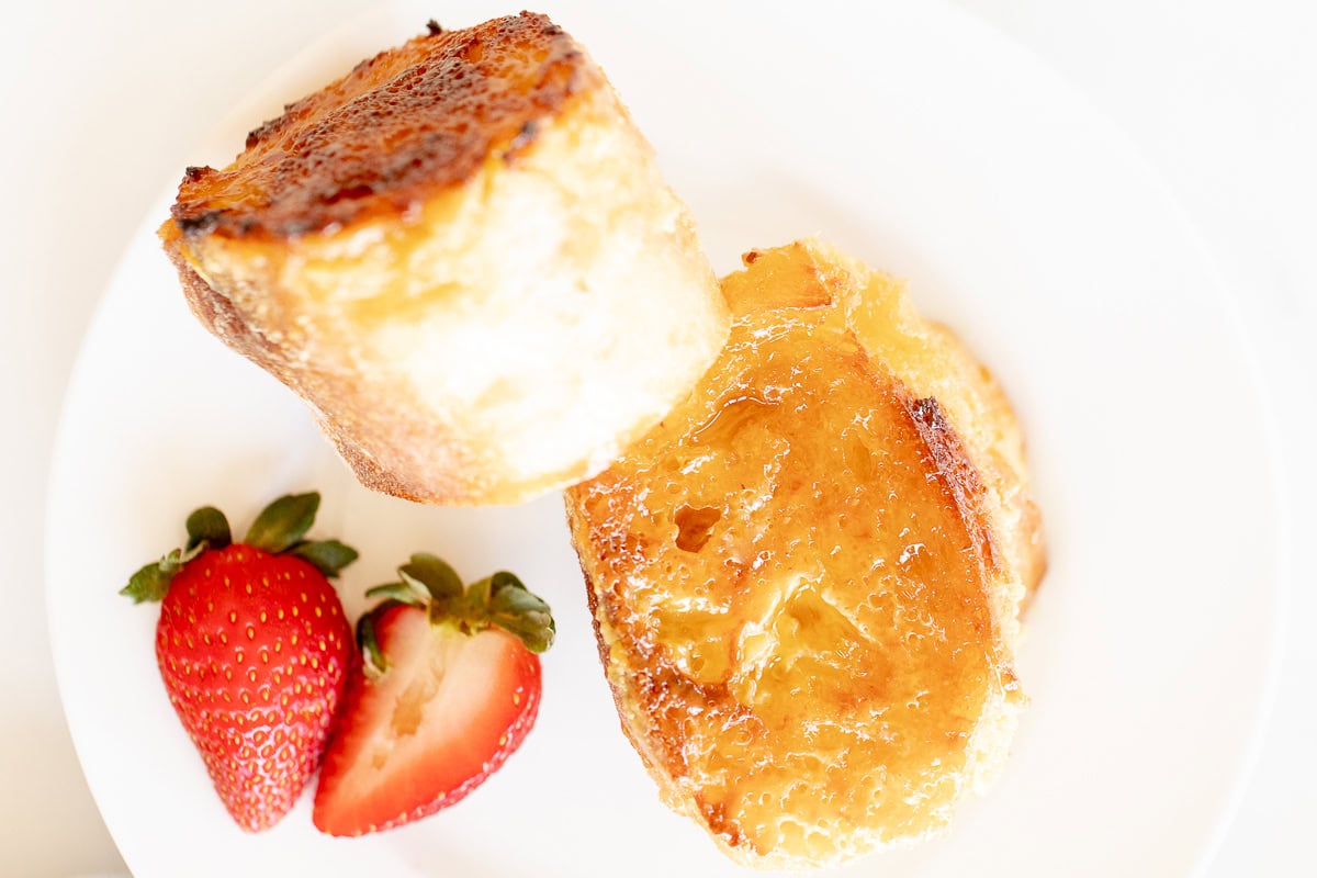 A plate with a slice of slice of French toast with creme brulee topping and a strawberry, served on a white background.