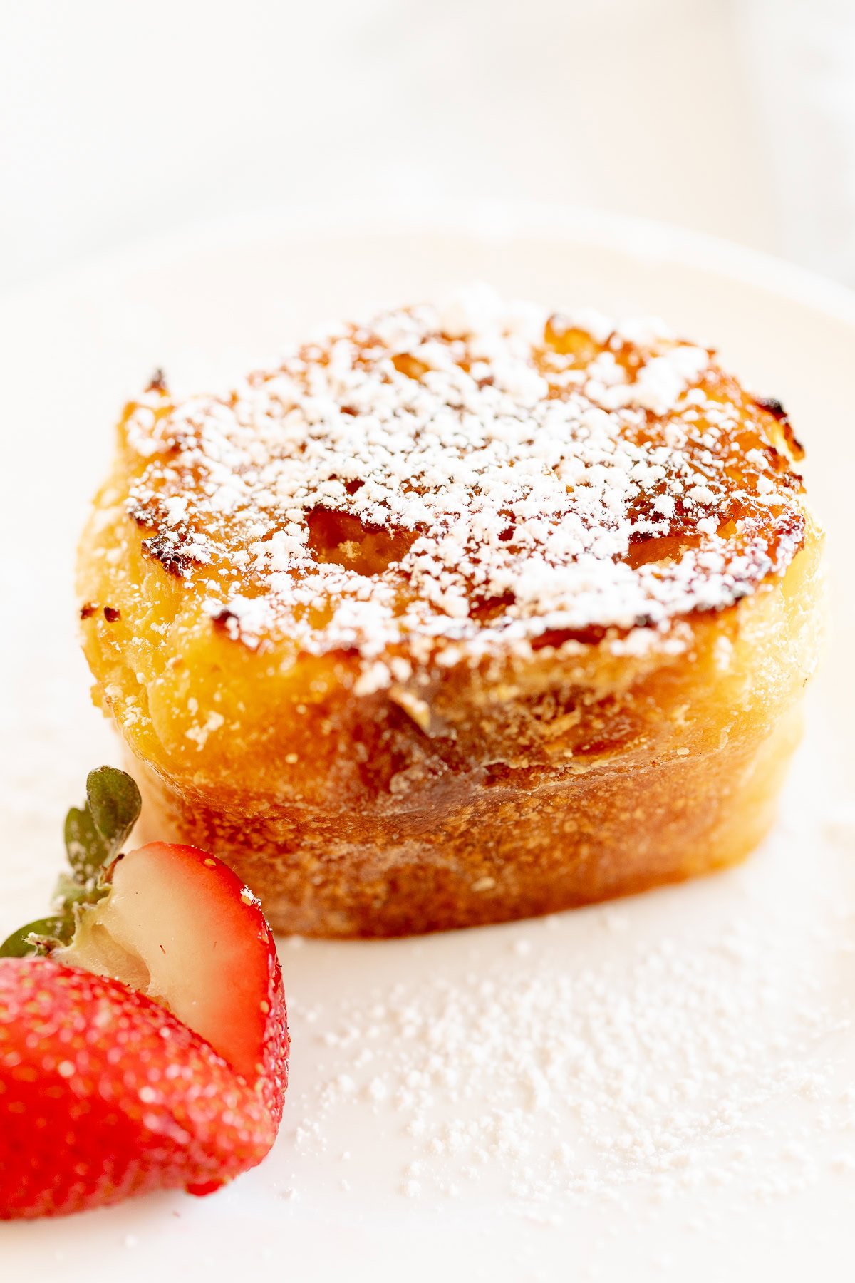 Creme Brulee French toast slice on a white plate with a gold fork. Garnished with sliced strawberries.