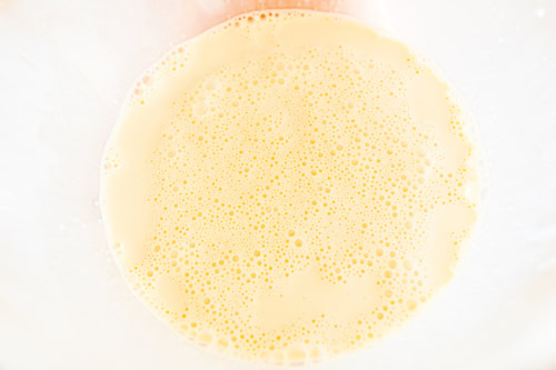 Close-up view of a bowl containing Crème Brûlée French Toast Casserole batter, showing a smooth, creamy texture with numerous small bubbles on the surface.