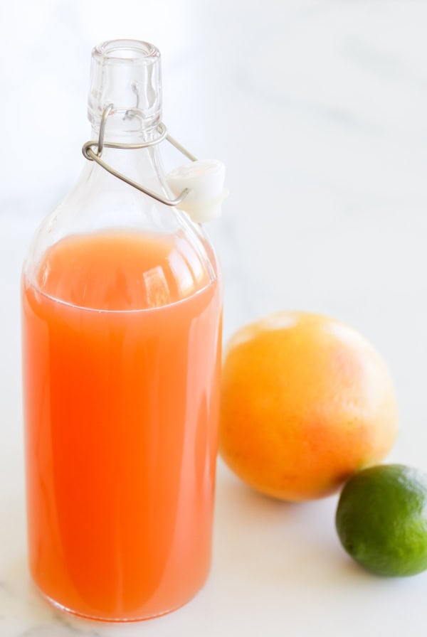 A glass bottle filled with orange juice, sealed with a ceramic stopper, next to a whole grapefruit and lime on a marble surface.