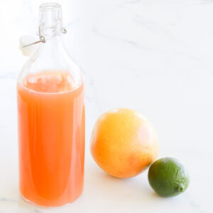 A clear glass bottle of fresh paloma mix, next to a grapefruit and lime on a marble surface.