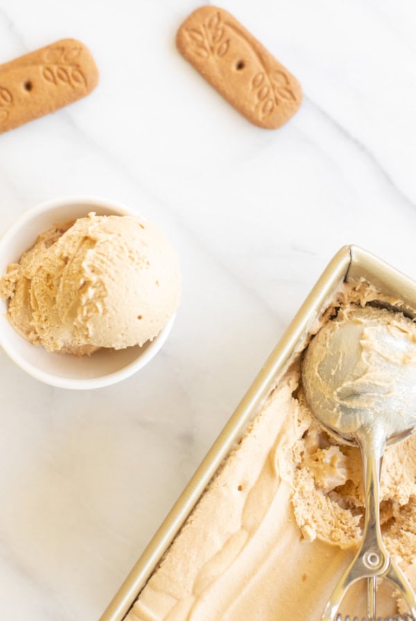 A scoop of cookie butter ice cream in a white bowl, with a container and a scooper, and cookies on a marble surface.