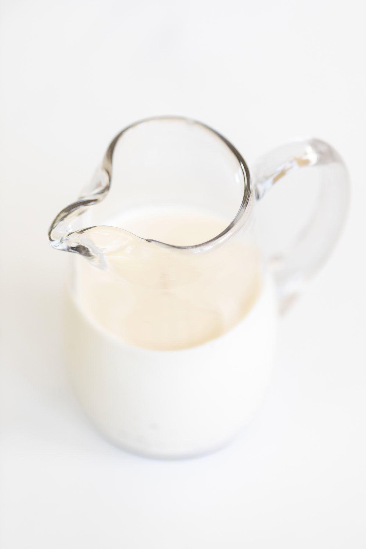 Clear glass pitcher filled with creamy pasta salad dressing on a white background.