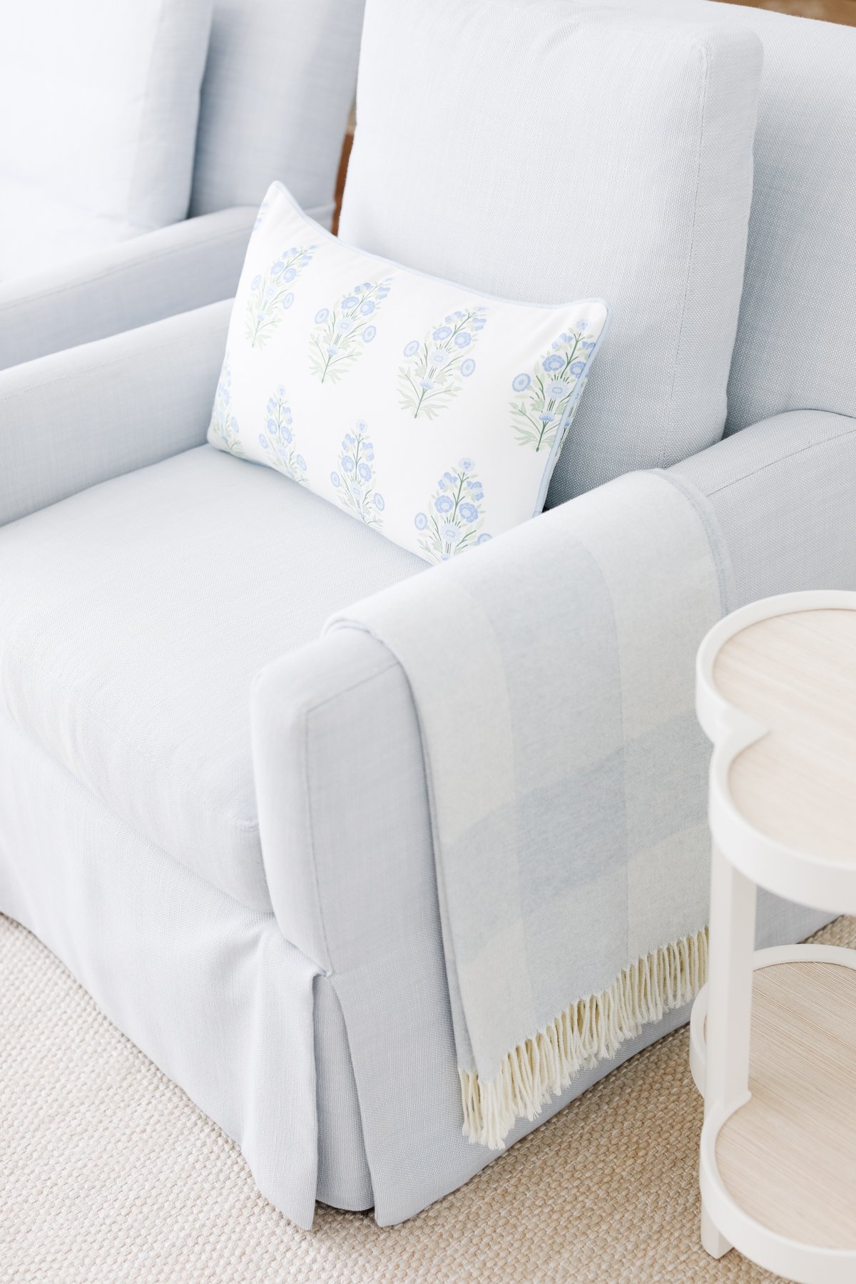 A light blue armchair with a floral print pillow and a checkered throw blanket draped over the arm, next to a small white side table.