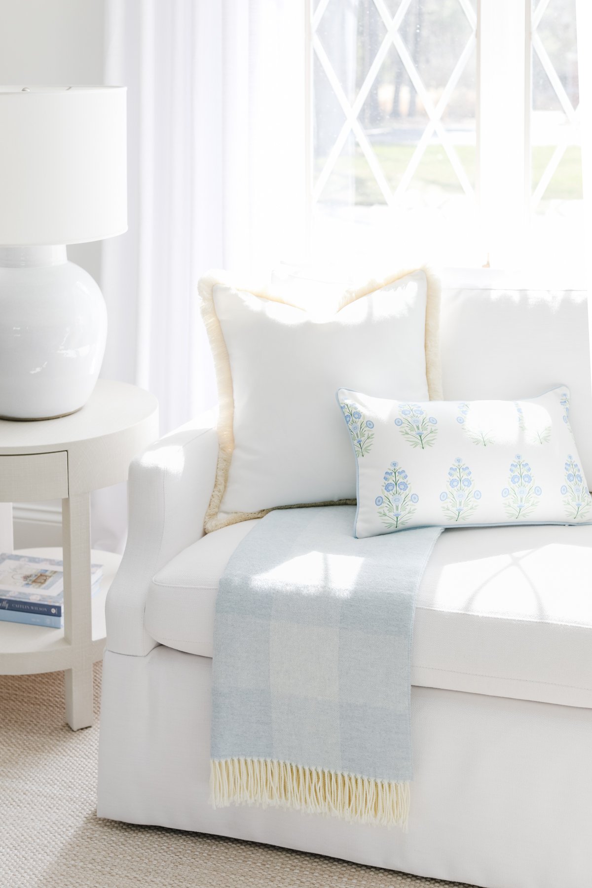 A white sofa with white and floral cushions is placed next to a small, round side table with a white lamp in a bright living room. A light blue throw blanket is draped over the sofa.