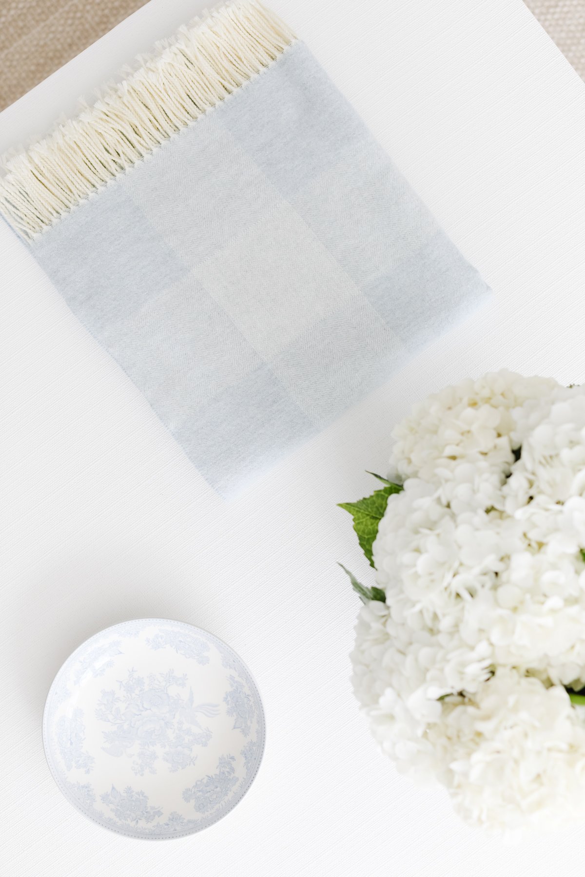 Top-down view of a light blue and white checkered throw blanket with fringes, a white and blue decorative plate, and a bouquet of white hydrangeas on a white surface.