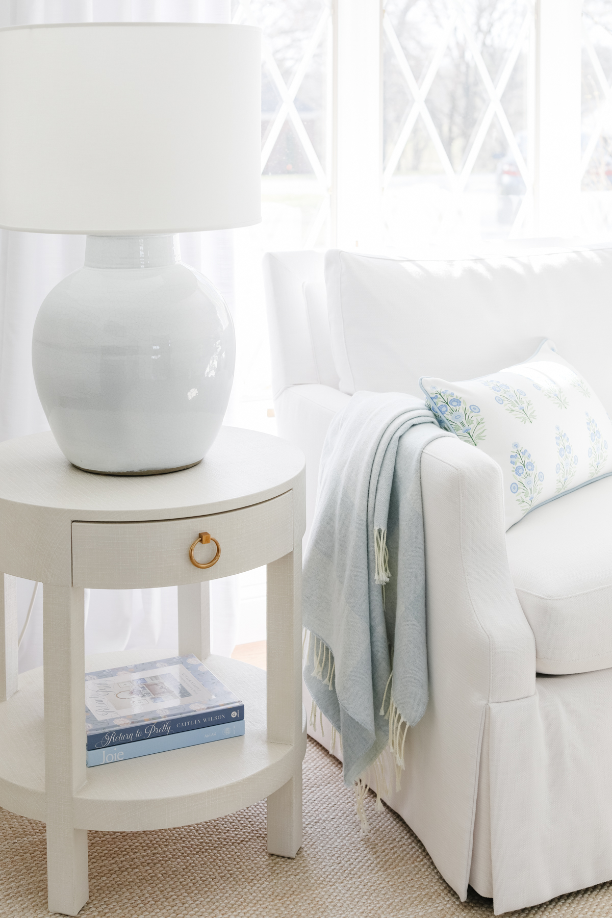 Side table with a white lamp sits next to a white sofa. Sofa has a light blue throw and floral pillow. Table holds books and is placed on a beige textured rug. Light streams in through diamond-paned windows.