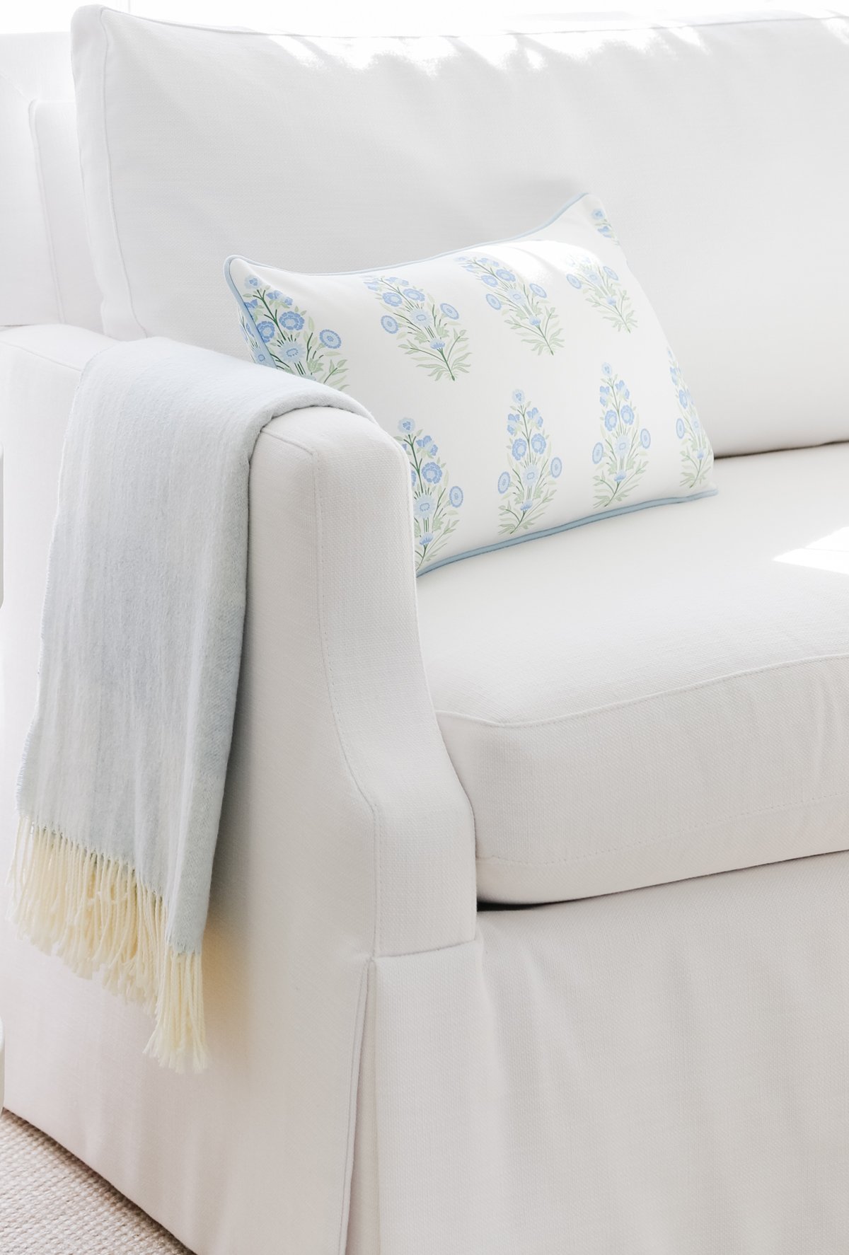 A white couch with a light blue blanket draped over the armrest and a white pillow decorated with a blue floral pattern placed on the seat.