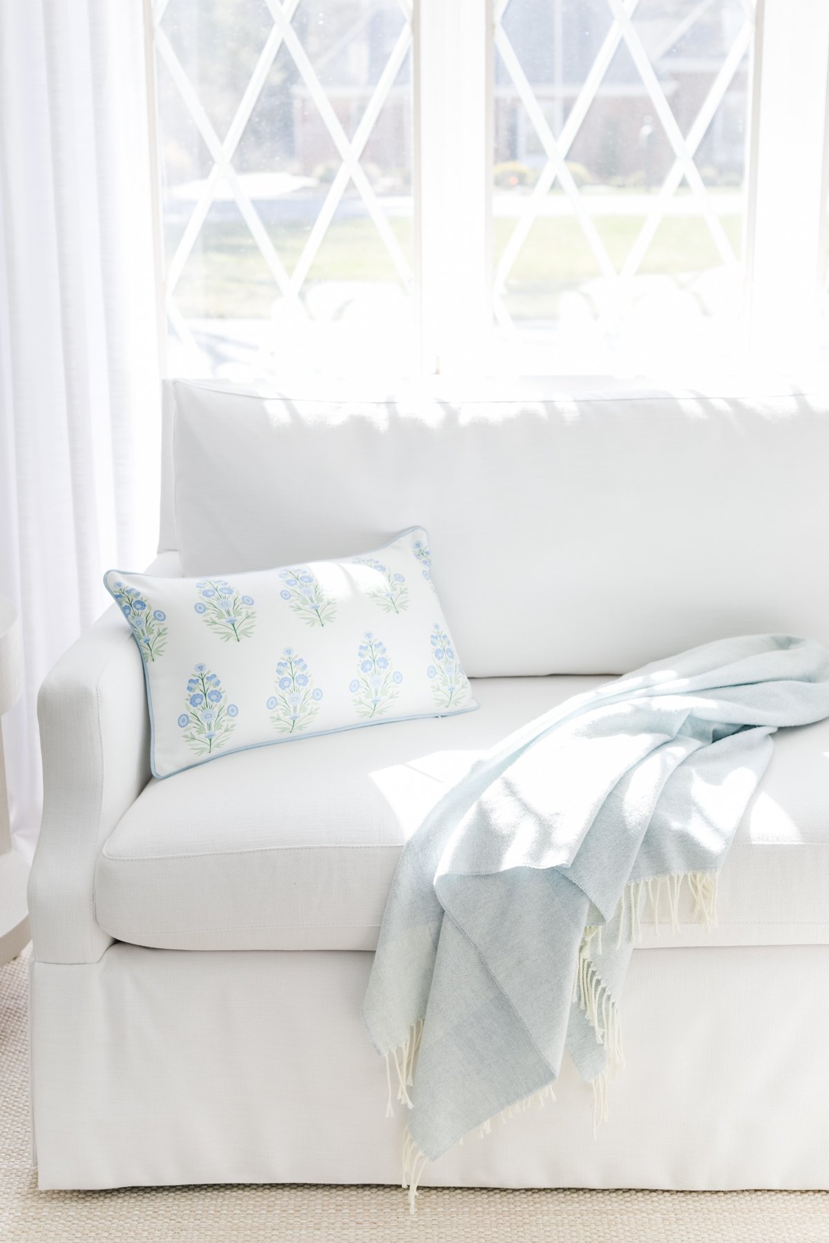 A white sofa with a floral cushion and a light blue throw blanket is positioned near a window with geometric patterns. Bright natural light fills the room.
