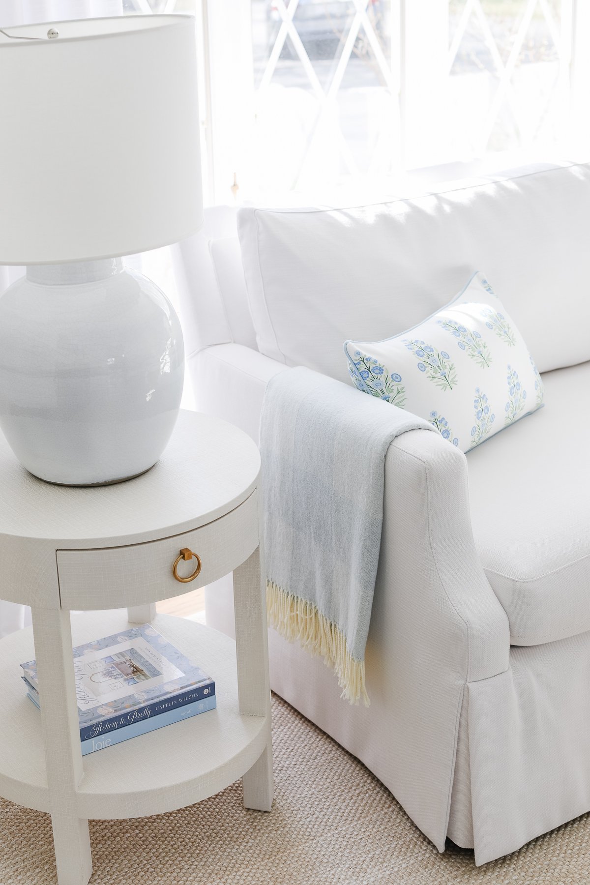 A white sofa with a floral pillow and blue throw blanket next to a round side table holding a white lamp and books.