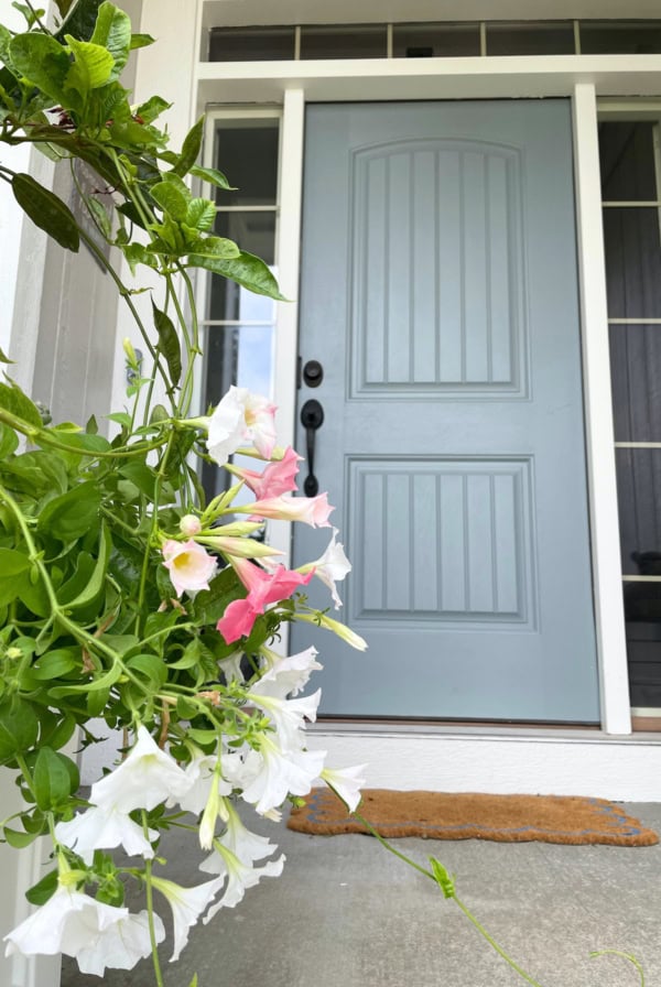 A light blue front door, painted in Benjamin Moore Brewster Gray, is framed by white and pink flowers on green vines. The door is set within a wooden porch, and a brown doormat lies in front of it.