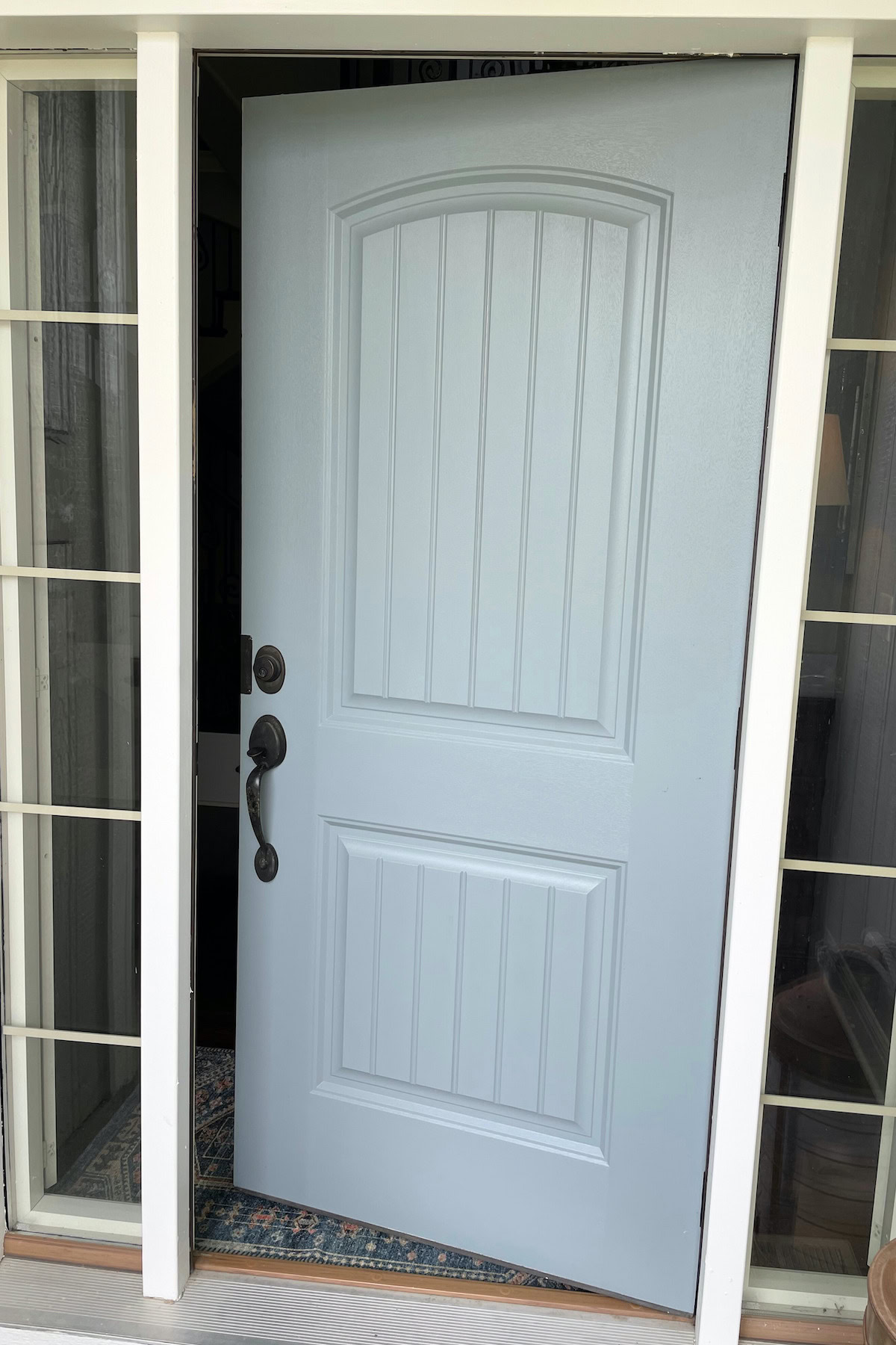 A partially open light blue front door painted in Benjamin Moore Brewster Gray with decorative paneling viewed head-on. It has a dark handle and is framed by glass windows on both sides.