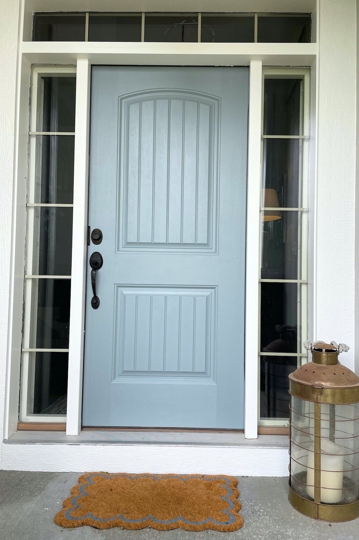 Front view of a blue door, painted in Benjamin Moore Brewster Gray, with a small glass window at the top, a black door handle and lock, flanked by vertical glass panes. A brown doormat sits in front, and a lantern is placed to the right.