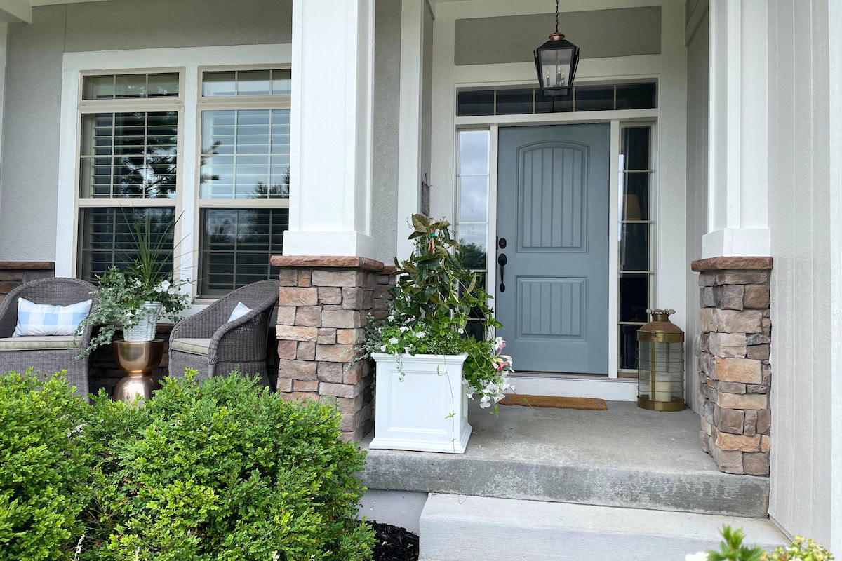Front porch with a Benjamin Moore Brewster Gray door, stone columns, and white trim. There are two wicker chairs with cushions, potted plants, and a lantern. Bushes and greenery surround the steps.