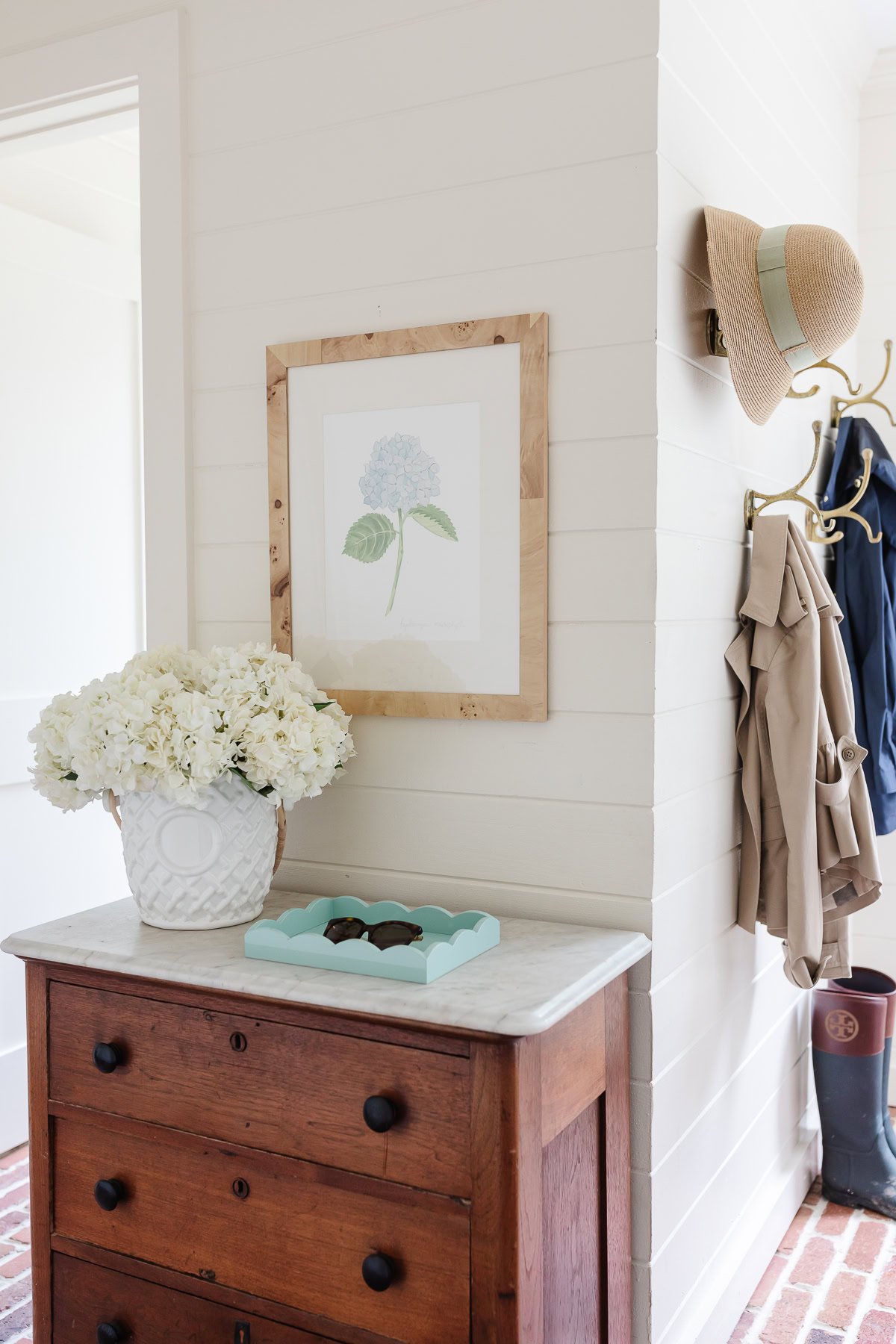 A wooden dresser with a white marble top holds a vase of flowers. Above it hangs a framed floral print on walls painted in a Benjamin Moore white color. On the wall are hanging coats, a hat, and rain boots. A light turquoise tray sits on the dresser.