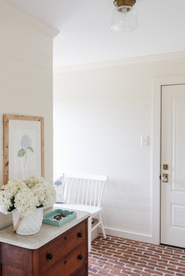 A bright entryway painted in a Benjamin Moore white color, with a brick floor, small wooden dresser holding a vase of white flowers, a framed flower print on the wall, a white bench with a pillow, and a closed white door.