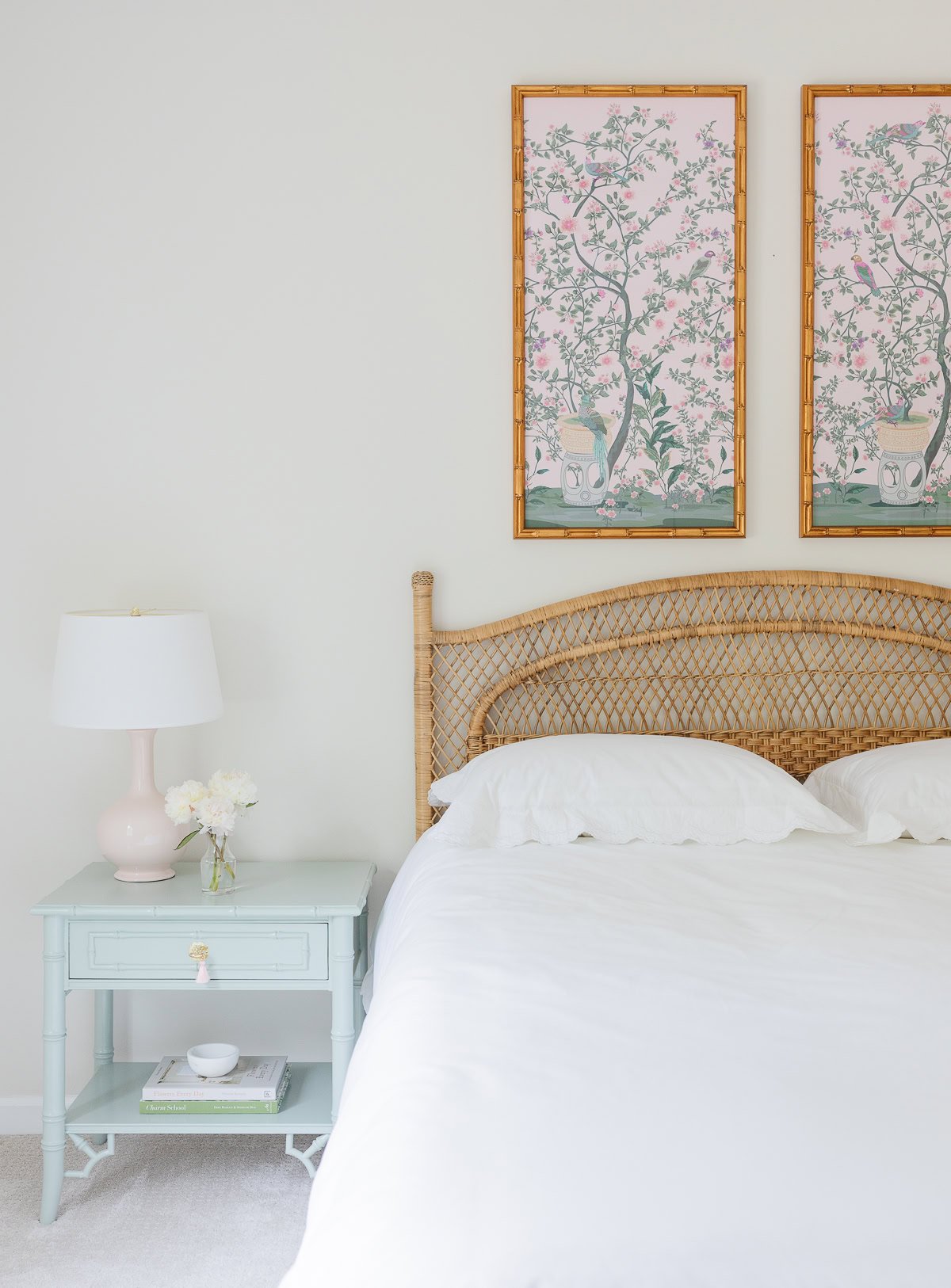 A bedroom features a rattan bed frame with walls painted in a Benjamin Moore White Color, a light blue nightstand with a lamp and flowers, and two framed botanical artworks on the wall.